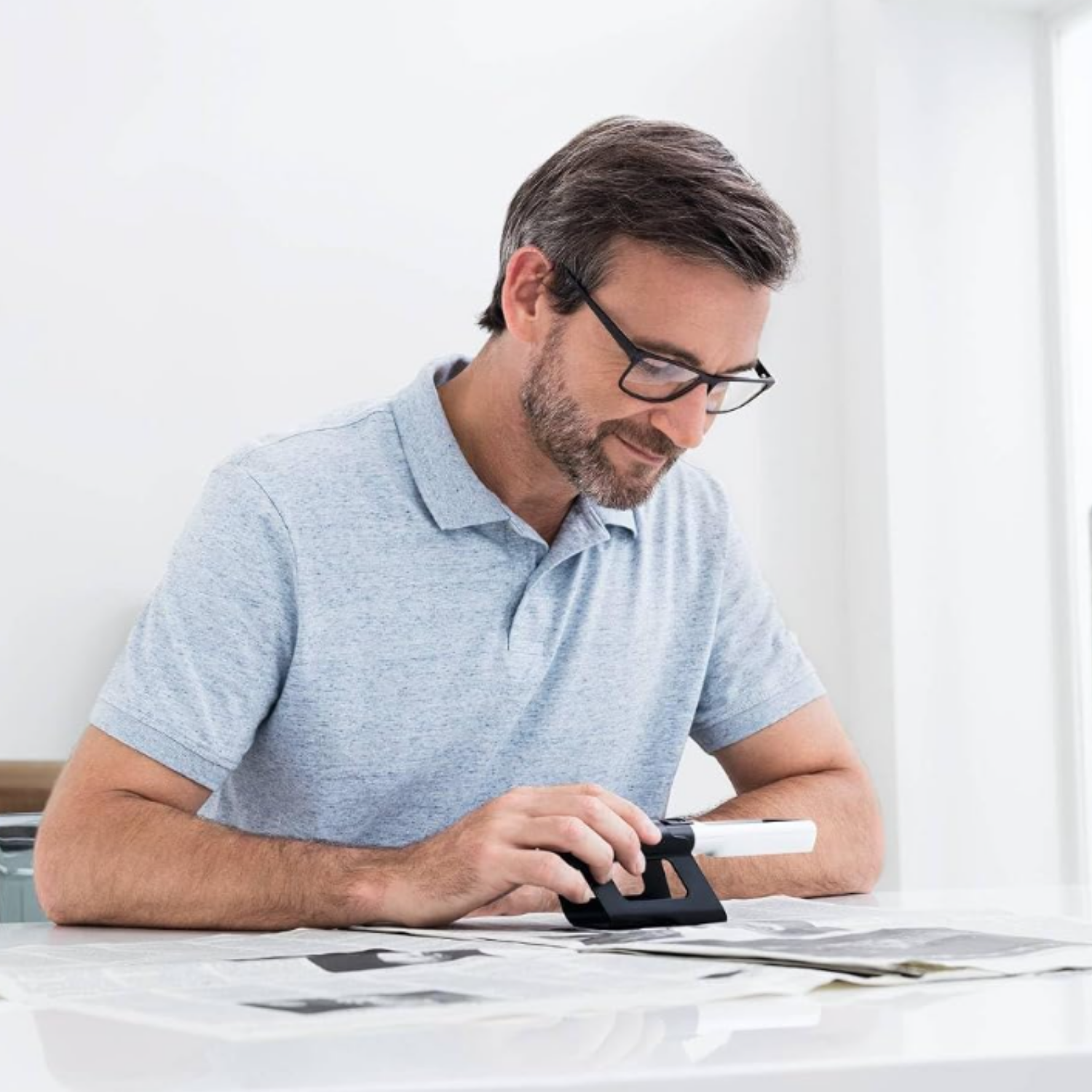 A man wearing a light blue polo shirt  sits at a white table, using a black magnifying glass in a stand.