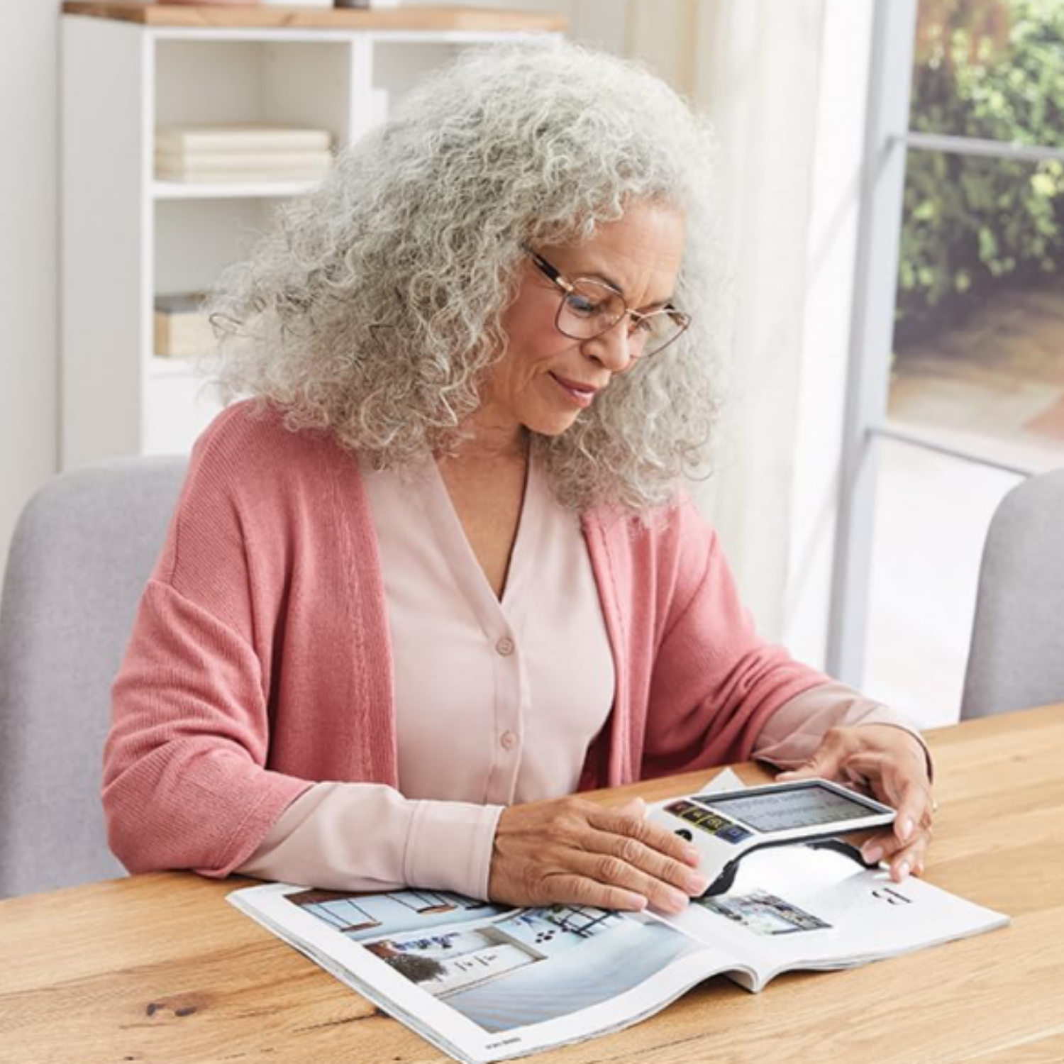 Older woman using handheld ideo magnifier to read magazine at a table.
