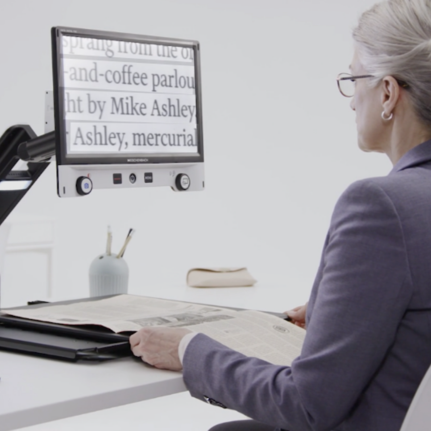 A woman in a purple blazer sits at a white desk, reading magnified text on the Vario Advanced CCTV.