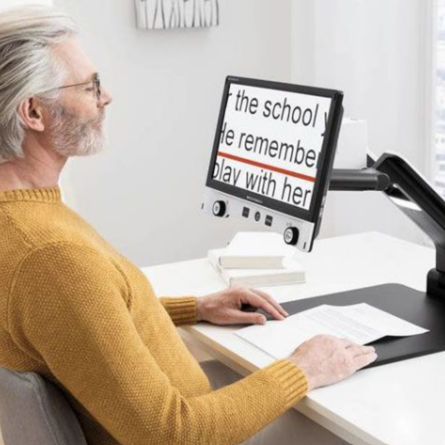 A man in a mustard yellow sweater sits at a desk using a desktop video magnifier to read enlarged text.